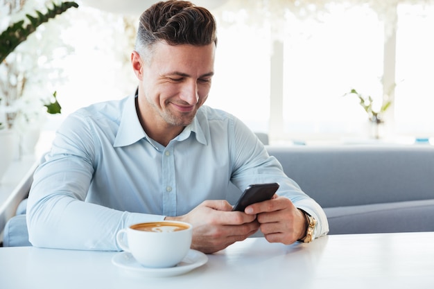 Imagem de alegre homem maduro sentado sozinho no café da cidade com uma xícara de cappuccino e digitando a mensagem de texto usando o celular preto