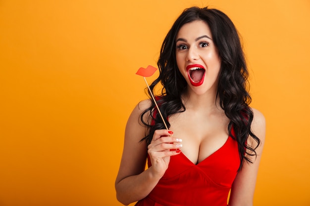 Imagem de alegre mulher 20 anos de vestido vermelho, sorrindo e segurando os lábios de papel na vara, isolado sobre a parede amarela