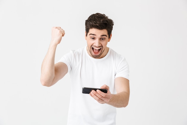 Imagem de animado feliz emocional jovem posando isolado sobre os jogos de parede branca pelo celular fazer gesto de vencedor.