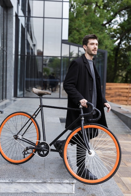 Imagem de bonito jovem empresário caminhando ao ar livre com uma bicicleta.