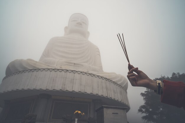 Imagem de Buda com incenso. Três incensos na mão de uma mulher contra um grande Buda no meio do nevoeiro. Colina Bana. Vietnã. Danang.