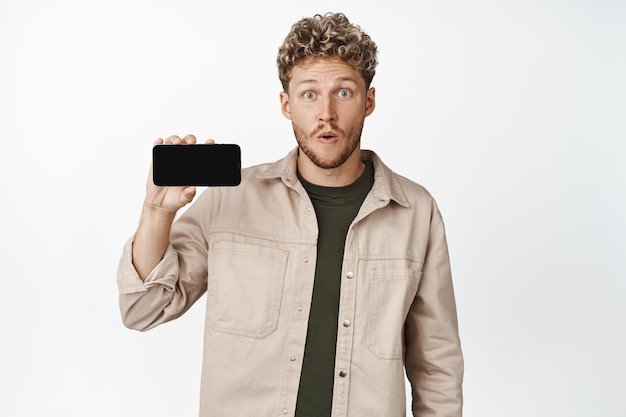 Imagem de cara espantado mostrando a tela do celular horizontalmente olhando impressionado para a câmera enquanto demonstra smth cool no fundo branco do aplicativo do smartphone