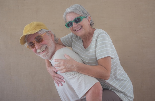 Imagem de casal sênior alegre com óculos de sol coloridos se divertindo enquanto homem pega carona em sua esposa isolada sobre fundo claro