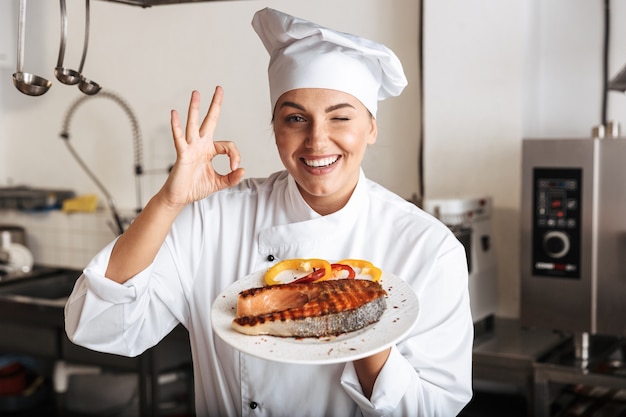 Imagem de chef de mulher feliz vestindo uniforme branco, segurando um prato com peixe grelhado na cozinha do restaurante