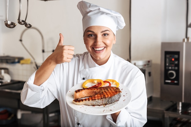 Imagem de chef mulher positiva vestindo uniforme branco, segurando um prato com peixe grelhado na cozinha do restaurante