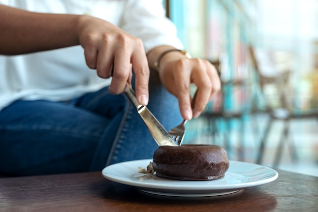 Imagem de close das mãos de uma mulher cortando um pedaço de donut de chocolate com uma faca e um garfo no café da manhã na mesa de madeira