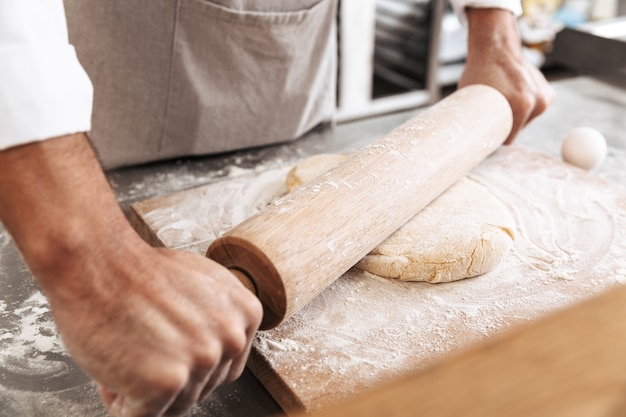 Imagem de close de mãos masculinas fazendo massa para pastelaria com rolo de madeira, na mesa da padaria ou cozinha