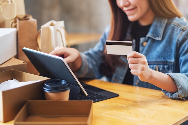 Imagem de close de uma jovem usando um tablet pc e um cartão de crédito para fazer compras online com uma caixa postal e sacolas de compras na mesa
