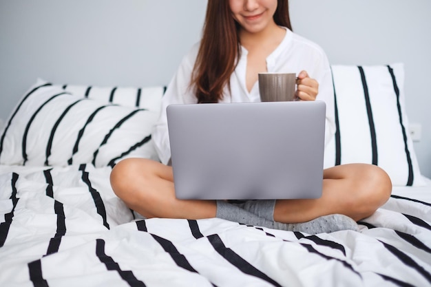 Imagem de close de uma linda mulher usando e trabalhando em um laptop, bebendo café enquanto está sentado em uma cama branca e aconchegante em casa