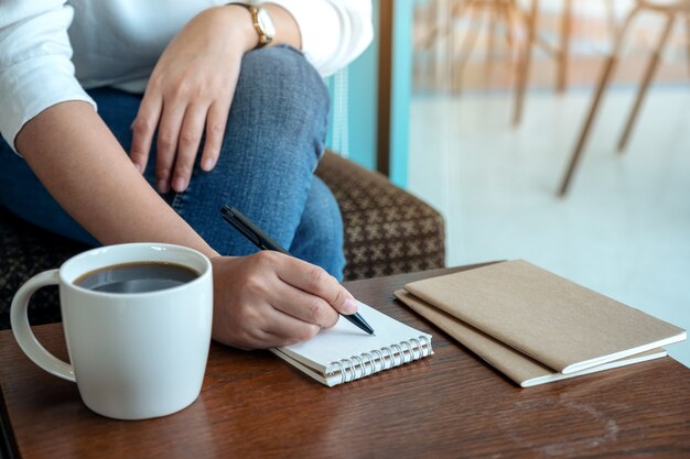 Imagem de close de uma mão de mulher escrevendo em um caderno em branco com uma xícara de café na mesa de madeira