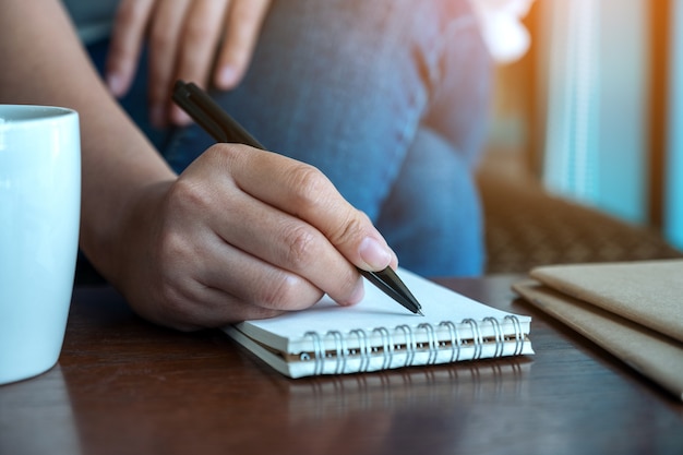 Imagem de close de uma mão de mulher escrevendo em um caderno em branco com uma xícara de café na mesa de madeira