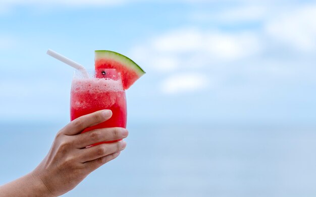 Imagem de close de uma mão segurando um copo de suco de melancia à beira-mar com o fundo do céu azul