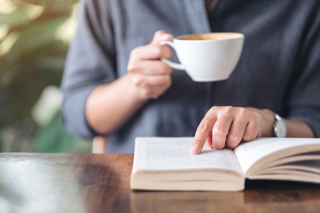 Imagem de close de uma mulher apontando e lendo um livro enquanto bebe café na mesa de madeira