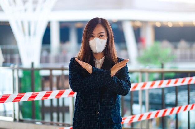 Imagem de close de uma mulher asiática usando máscara protetora, fazendo sinais de braços cruzados na frente da área da fita vermelha e branca para prevenir a propagação do conceito de pandemia covid-19