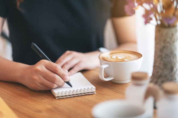 Imagem de close de uma mulher escrevendo em um caderno em branco com uma xícara de café na mesa de madeira