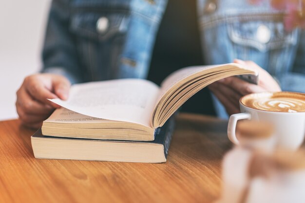 Imagem de close de uma mulher segurando e lendo um livro de romance vintage com uma xícara de café na mesa de madeira