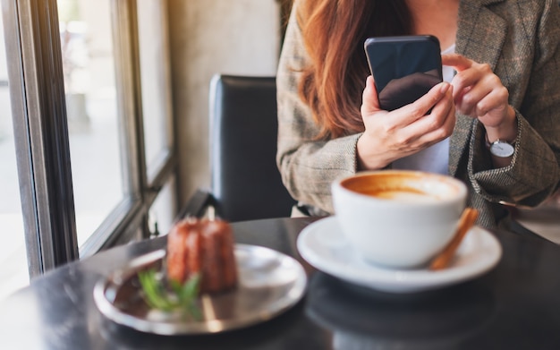 Imagem de close de uma mulher segurando e usando um telefone celular com uma xícara de café e um lanche na mesa no café