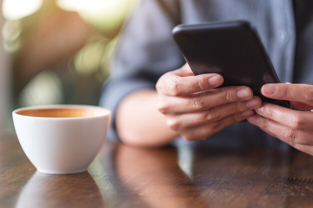 Imagem de close de uma mulher segurando e usando um telefone celular com uma xícara de café na mesa de madeira em um café