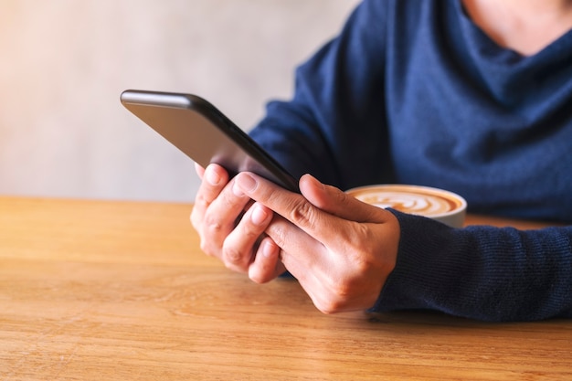 Imagem de close de uma mulher segurando e usando um telefone celular com uma xícara de café na mesa de madeira