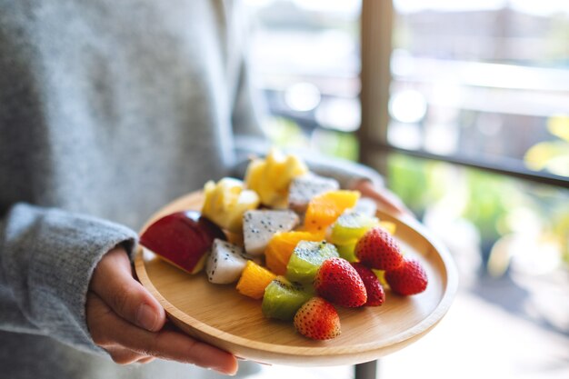 Imagem de close de uma mulher segurando um prato de madeira com frutas frescas em espetos