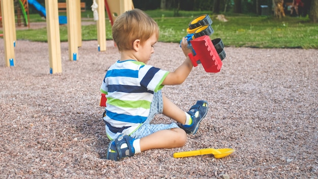 Imagem de close up de menino bonitinho brincando no palyground com brinquedos. Criança se divertindo com caminhão, escavadeira e reboque