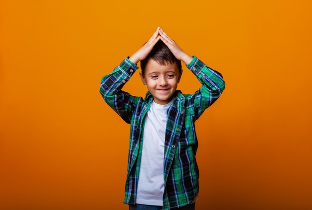 Imagem de close-up de um menino com as duas mãos acima da cabeça e fazendo um gesto no telhado, sobre um fundo amarelo.