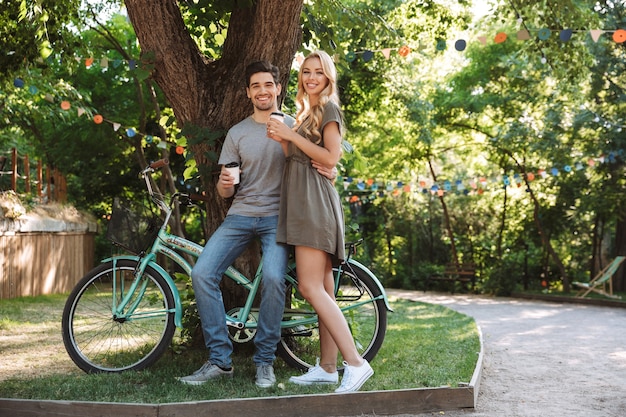 Imagem de corpo inteiro de alegre casal jovem posando junto perto de uma bicicleta e olhando para a câmera com café ao ar livre