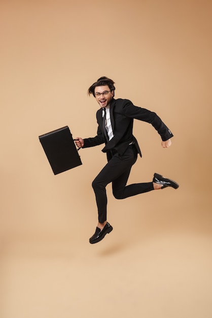 Imagem de corpo inteiro de um empresário caucasiano em um terno formal correndo com uma pasta preta isolada na parede bege