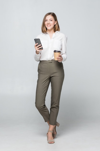 Imagem de corpo inteiro de uma bela mulher de negócios com roupa formal em pé e usando o telefone celular com café para viagem na mão isolado sobre o cinza