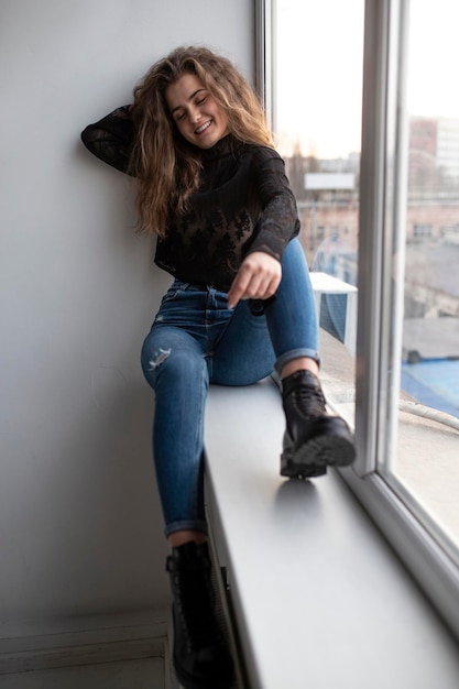 Imagem de corpo inteiro de uma linda jovem alegre com cabelo encaracolado, vestindo blusa preta e jeans posando perto de janelas