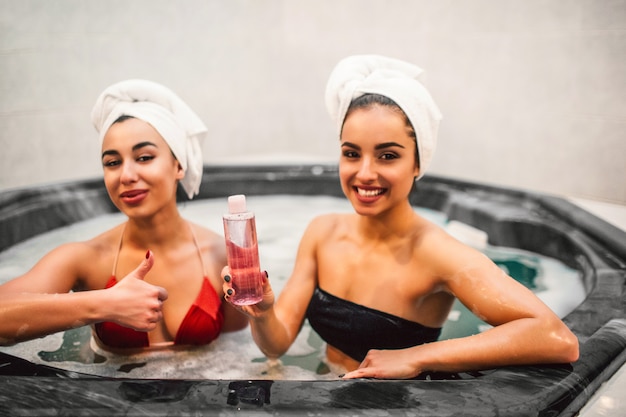 Imagem de dois modelos sentados no banho de hidromassagem cheio de água com espuma. Odel Europeu segurar gel de banho nas mãos. Mulher asiática espera grande polegar. Eles parecem retos.
