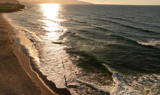 Imagem de drone de pessoas em uma praia de areia banhada pelas ondas espumosas do mar sob o sol forte ao pôr do sol