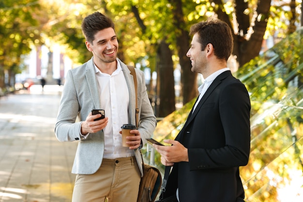 Imagem de empreendedores masculinos de terno usando smartphone enquanto caminhava ao ar livre pelo parque verde com café para viagem, em dia de sol