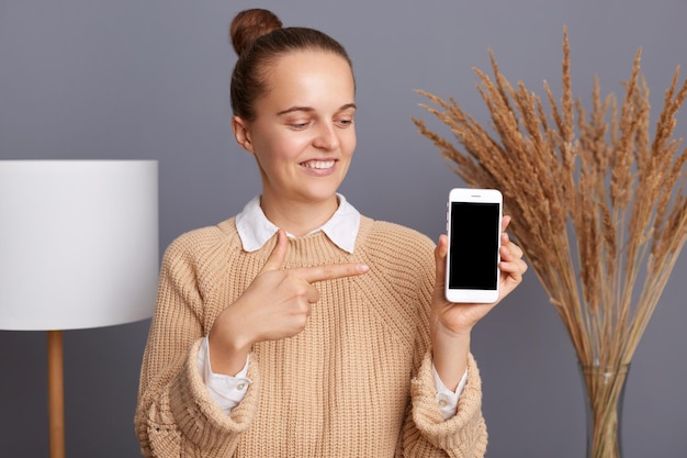 Foto imagem de encantadora jovem adulta caucasiana bonita vestindo jumper bege em pé segurando o telefone inteligente apontando o dedo na tela vazia para anúncio ou texto promocional