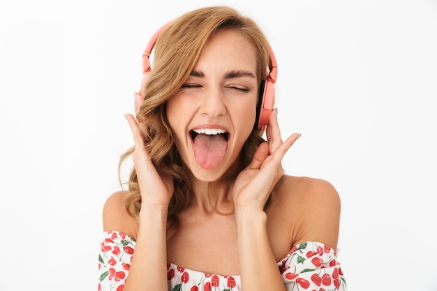 Imagem de engraçado feliz jovem posando isolado sobre uma parede branca, ouvindo música com fones de ouvido, mostrando a língua.