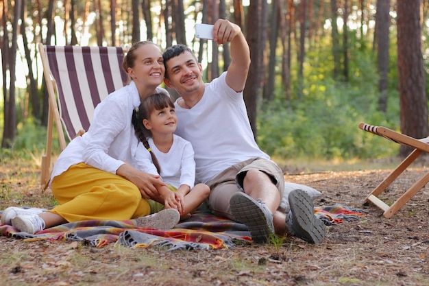 Imagem de feliz e positiva família marido esposa e filha sentada no chão no cobertor e tirando selfie de suas férias na floresta sorrindo alegremente