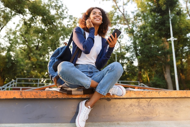 Imagem de feliz jovem africana sentada ao ar livre no parque, usando telefone celular, ouvindo música com fones de ouvido.