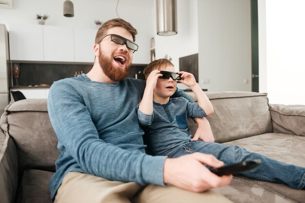 Imagem de feliz pai barbudo segurando o controle remoto enquanto assiste tv com seu filho pequeno usando óculos 3d.