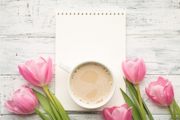 Imagem de foto de vista superior de perto do bloco de notas em branco de papel aberto com uma xícara de cappuccino de espuma e tulipas rosa pastel isoladas mesa de mesa de madeira branca