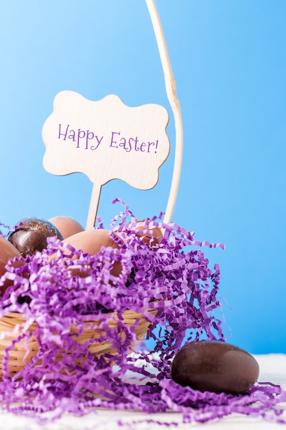 Imagem de frango, ovos de chocolate e papel decorativo roxo em uma cesta na parede azul vazia com um desejo de feliz Páscoa