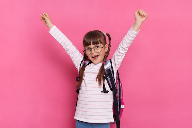 Imagem de garotinha vestindo camisa e óculos em pé com mochila isolada sobre fundo rosa levantou os braços gritando com expressão facial feliz