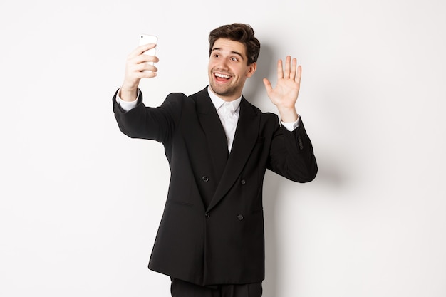 Imagem de homem bonito de terno, tendo uma videochamada e acenando com a mão para a câmera do smartphone, gravando um vídeo, cumprimentando alguém, em pé contra um fundo branco.