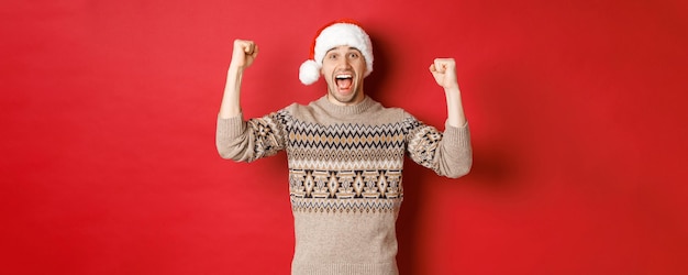 Imagem de homem bonito e alegre com chapéu de xerife e Papai Noel, comemorando o ano novo, triunfando ou ganhando algo, levantando as mãos e gritando de alegria, em pé sobre um fundo vermelho