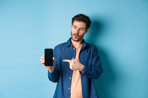 Imagem de homem bonito, falando sobre o aplicativo, apontando o dedo para a tela do celular e em pé sobre um fundo azul.
