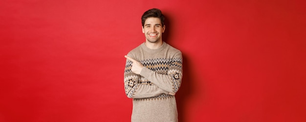 Imagem de homem bonito na camisola de natal, comemorando feriados de ano novo, sorrindo feliz e apontando o dedo no espaço de cópia do canto superior esquerdo, de pé sobre fundo vermelho.