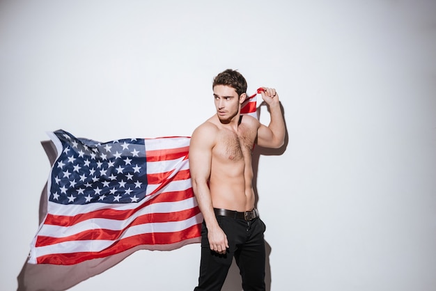 Imagem de homem bonito posando e desviar o olhar, segurando a bandeira dos EUA. Isolado sobre a parede branca.