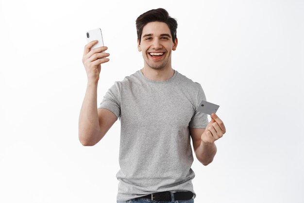 Imagem de homem caucasiano mostrando smartphone e cartão de crédito plástico pagando e fazendo compras online na internet sobre fundo branco