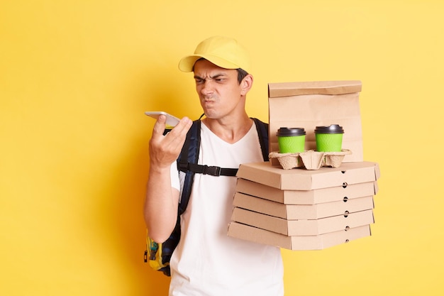 Imagem de homem de correio com raiva de boné amarelo e camiseta segurando pizza e copos de café usando celular para se conectar com o cliente posando isolado sobre fundo amarelo