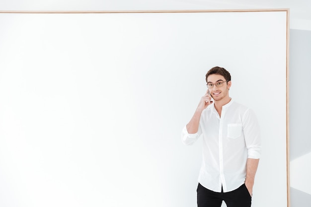 Imagem de homem feliz usando óculos e vestido com camisa branca, falando por telefone em pé perto do grande quadro. Olhe para o lado.