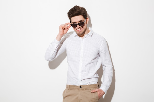 Imagem de homem feliz, vestido de camisa, olhando para a câmera debaixo de óculos de sol pretos e sorrindo mostrando dentes perfeitos, sobre espaço em branco com sombra